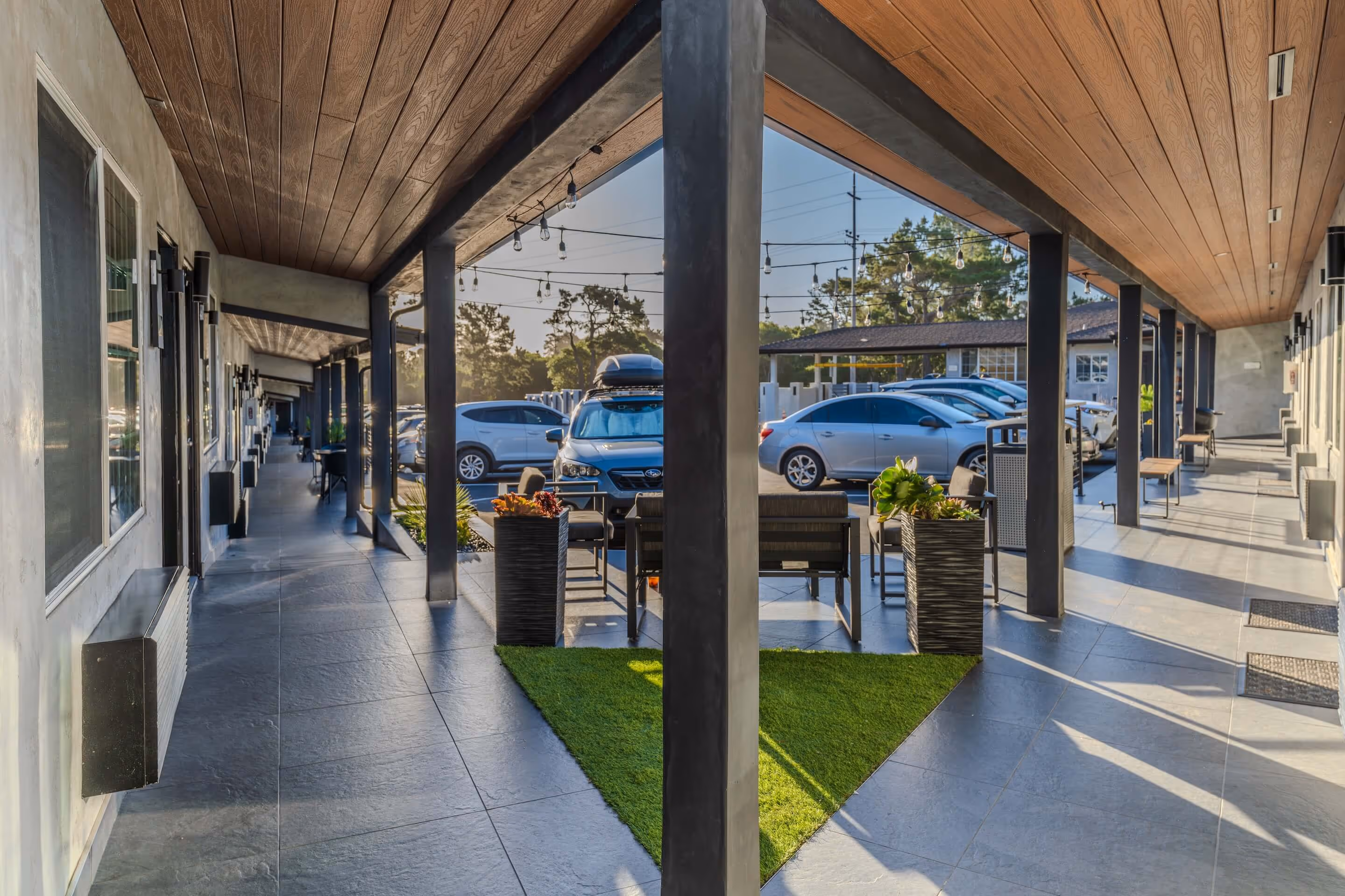 Outdoor corridor of a modern motel with parked cars, seating area with chairs and tables, and decorative string lights overhead.