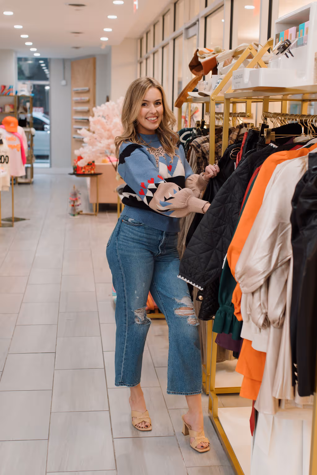 Smiling woman in a patterned sweater and ripped jeans browsing clothes on a rack in a brightly lit boutique store.