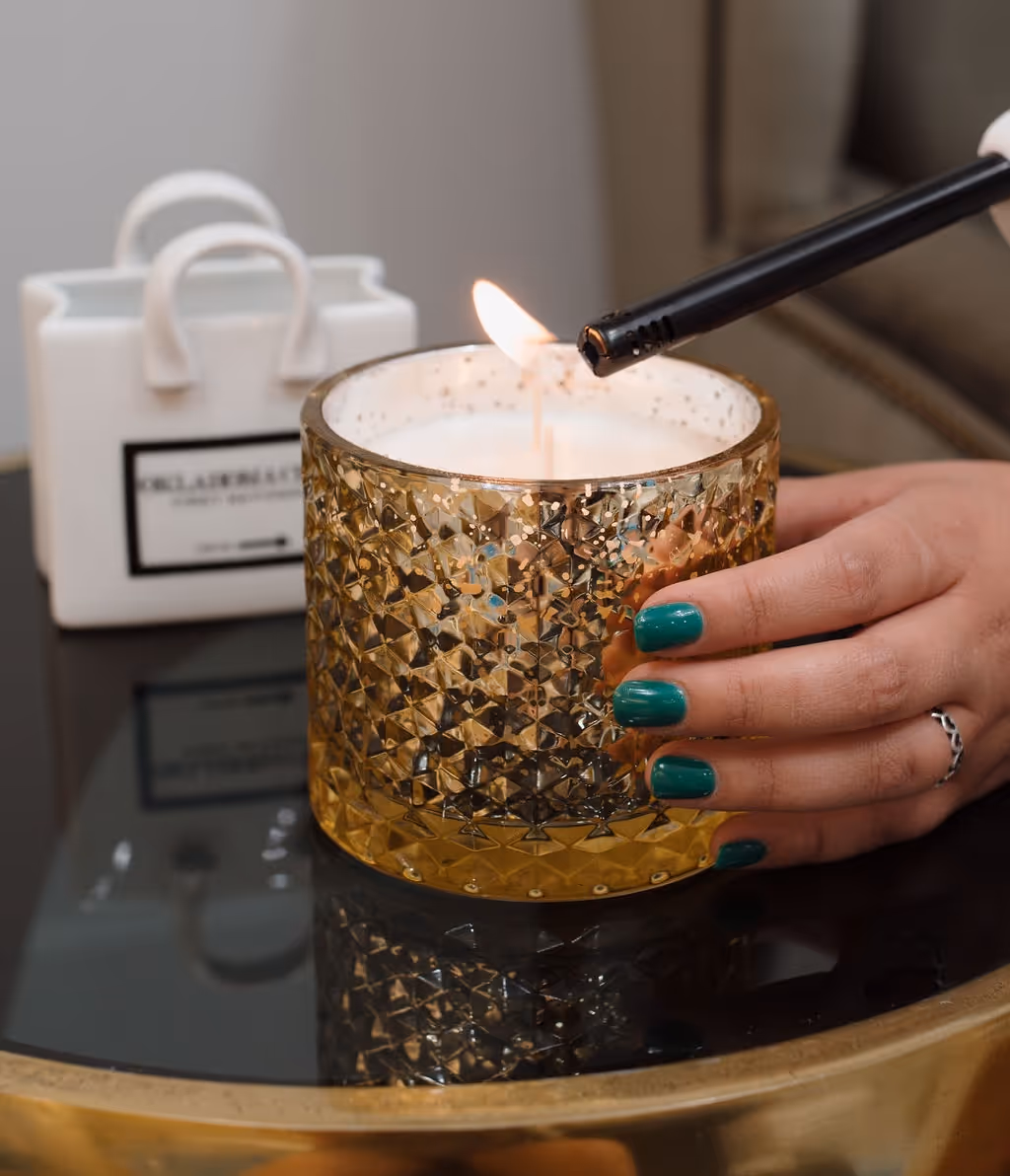 Hand with green nail polish lighting a candle in a textured golden glass holder on a reflective surface.