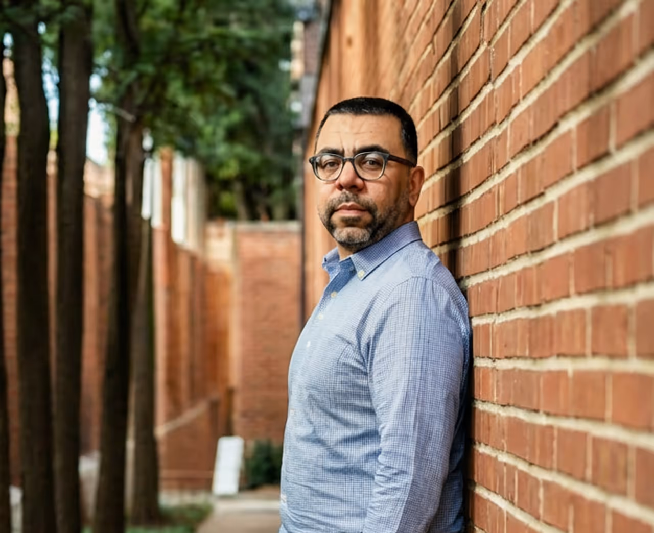 Man wearing glasses and a blue checkered shirt leaning against a brick wall with trees in the background.