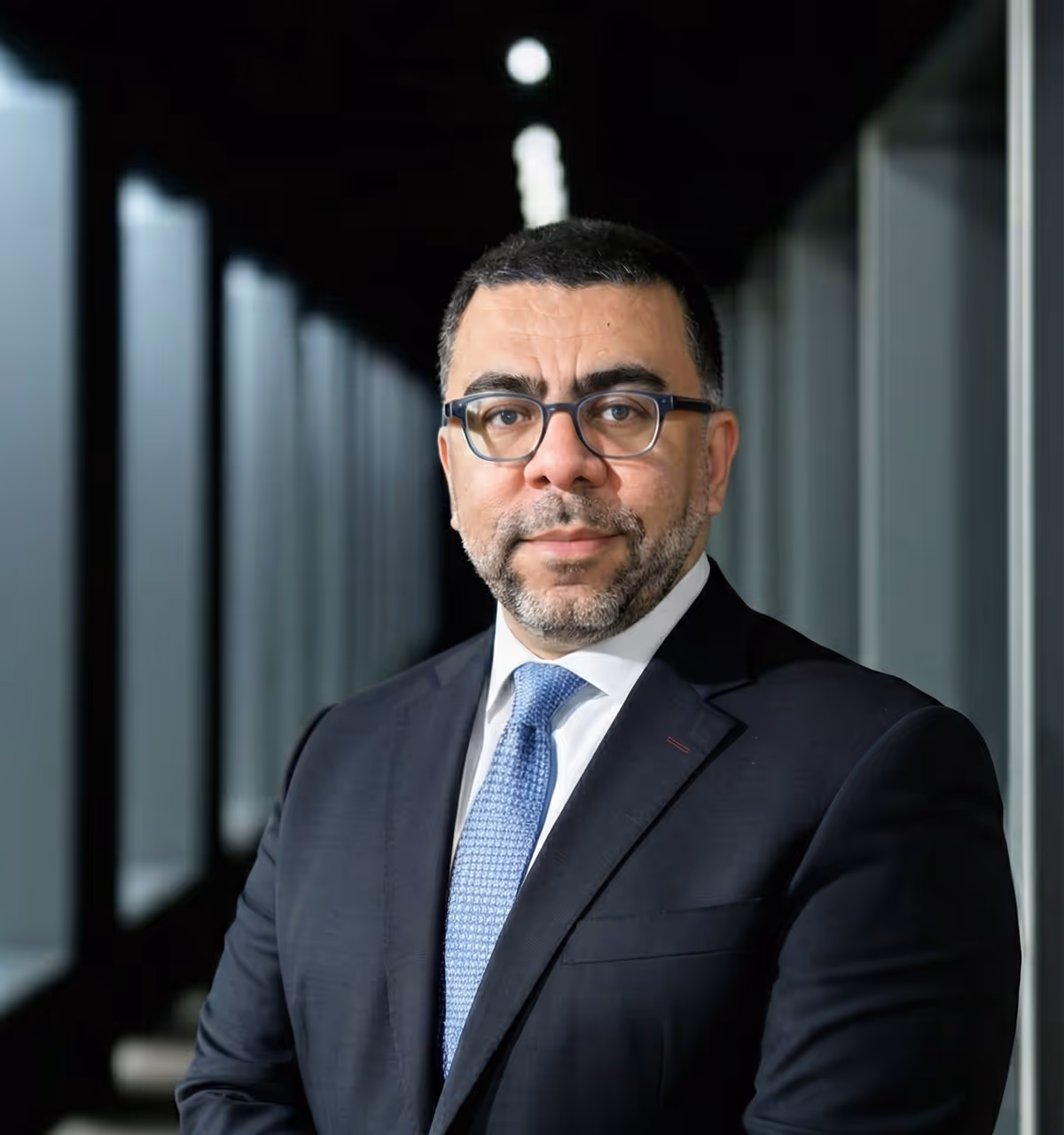Man with glasses wearing a dark suit and light blue tie standing in a modern corridor with glass walls.