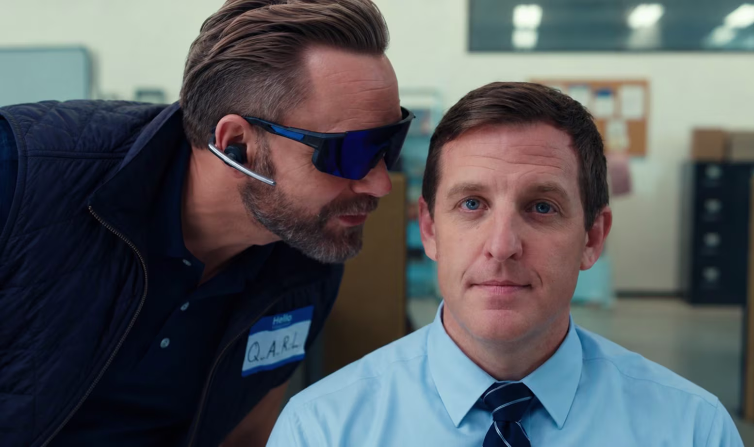 Q.A.R.L., in wraparound sunglasses, leans in to whisper to an auditor in a shirt and tie