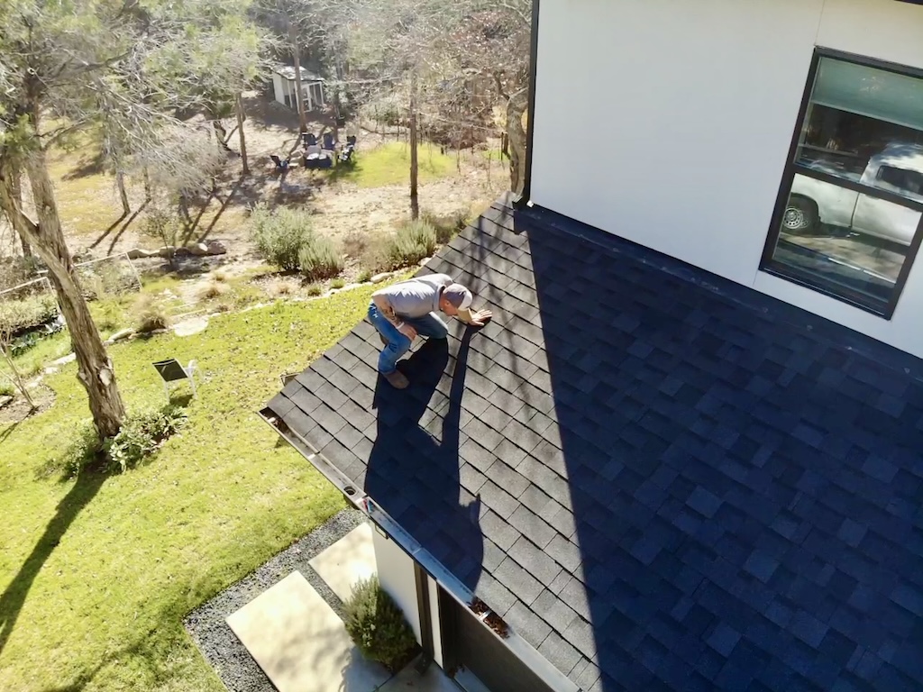 Man inspecting or repairing a dark-shingled roof of a white house on a sunny day with trees and lawn in the background.