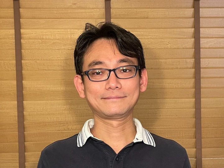Smiling man with black hair and glasses wearing a black collared shirt stands in front of a wooden backdrop.