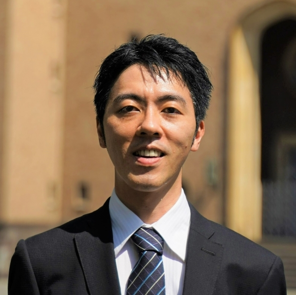 Smiling man in a black suit, white shirt, and striped tie standing outdoors with blurred building background.