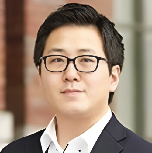 Smiling man with black hair and glasses wearing a black collared shirt stands in front of a wooden backdrop.