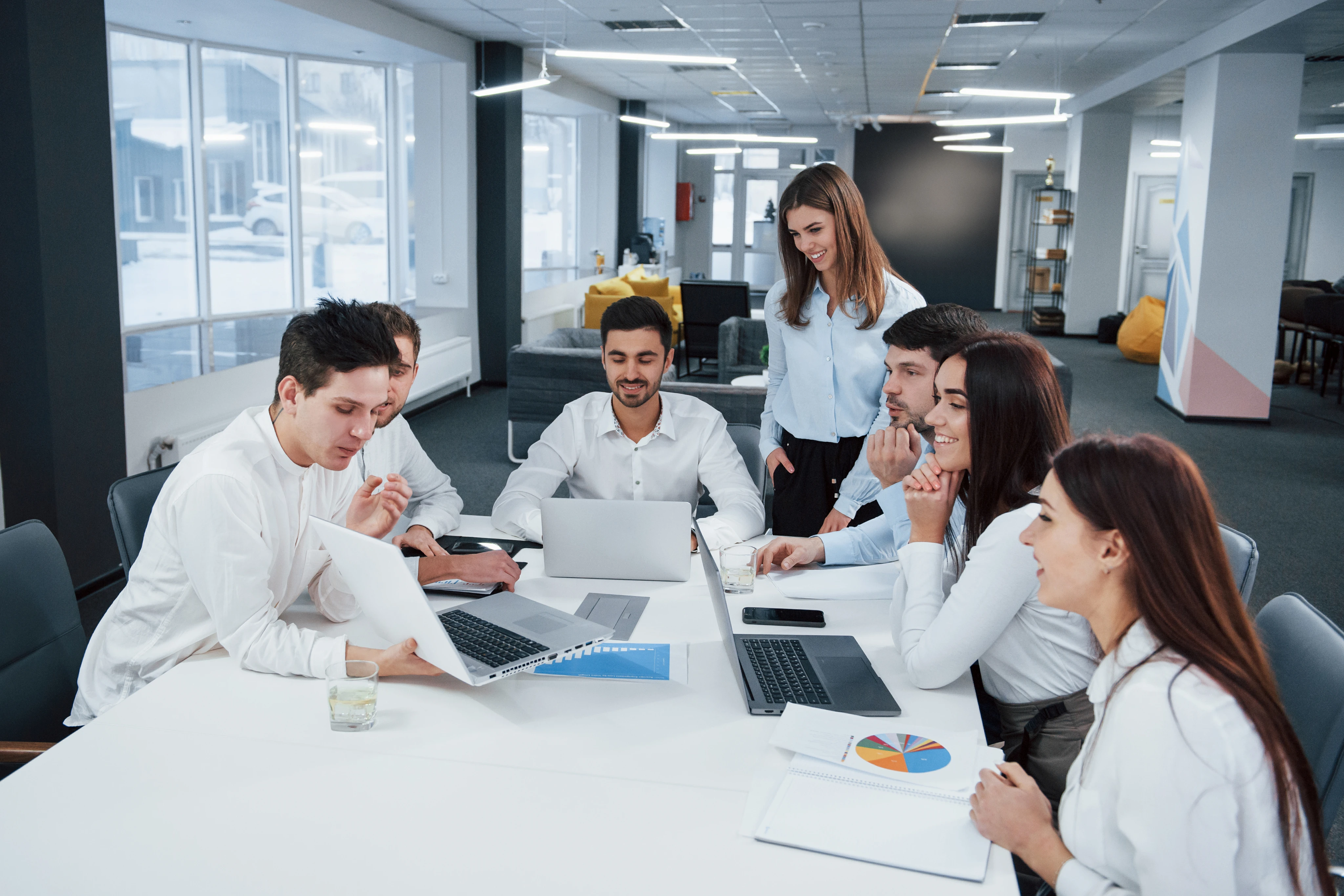A group of six young professionals gathered around a table in a modern office, collaborating with laptops and documents.