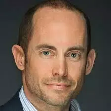 Portrait of a man with short brown hair and green eyes wearing a light blue shirt and dark jacket.