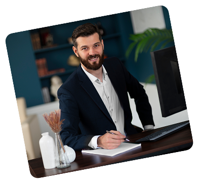 Smiling bearded man in a suit sitting at a desk with a computer and writing in a notebook.