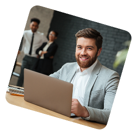 Smiling businessman in a gray blazer working on a laptop at a desk with two colleagues blurred in the background.