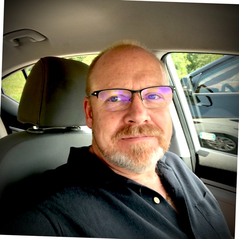 Middle-aged man with glasses and a beard sitting in the driver's seat of a car, smiling slightly.