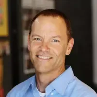 Smiling man with short brown hair wearing a light blue collared shirt.