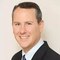 Smiling man with short dark hair wearing a dark suit, white shirt, and striped tie against a plain light background.