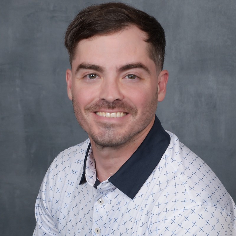 Smiling man with short brown hair and trimmed beard wearing a white shirt with blue crosses and a dark blue collar.