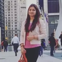 Woman with long dark hair wearing a pink top and light jacket standing outdoors in an urban area with buildings and people in the background.