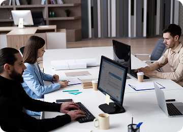 Three professionals working on computers at a white office table with documents and coffee cups.