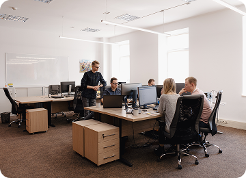 Four colleagues in an office collaborate around a desk with computers and laptops.
