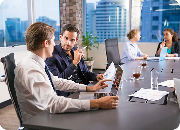 Two men discussing documents and laptop at a conference table while two women talk in the background in a modern office.