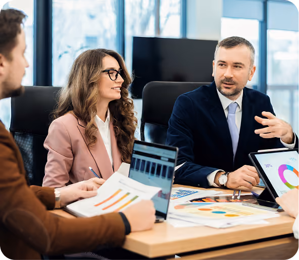 Three business professionals in a meeting room discussing charts and graphs displayed on laptops and printed papers.