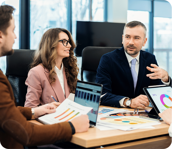 Three business professionals in a meeting room discussing charts and graphs displayed on laptops and printed papers.