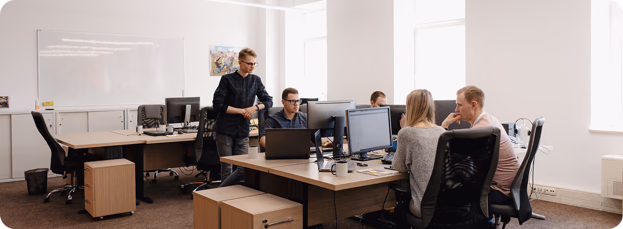 Five coworkers collaborating in a modern office around desks with computers and laptops.