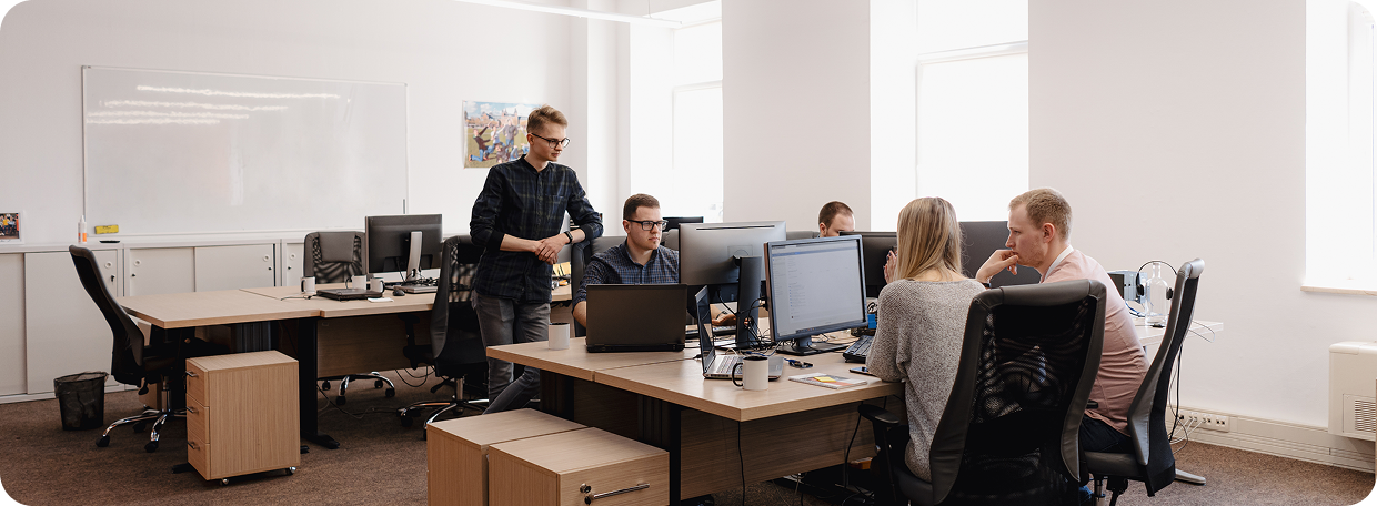 Five coworkers collaborating in a modern office around desks with computers and laptops.