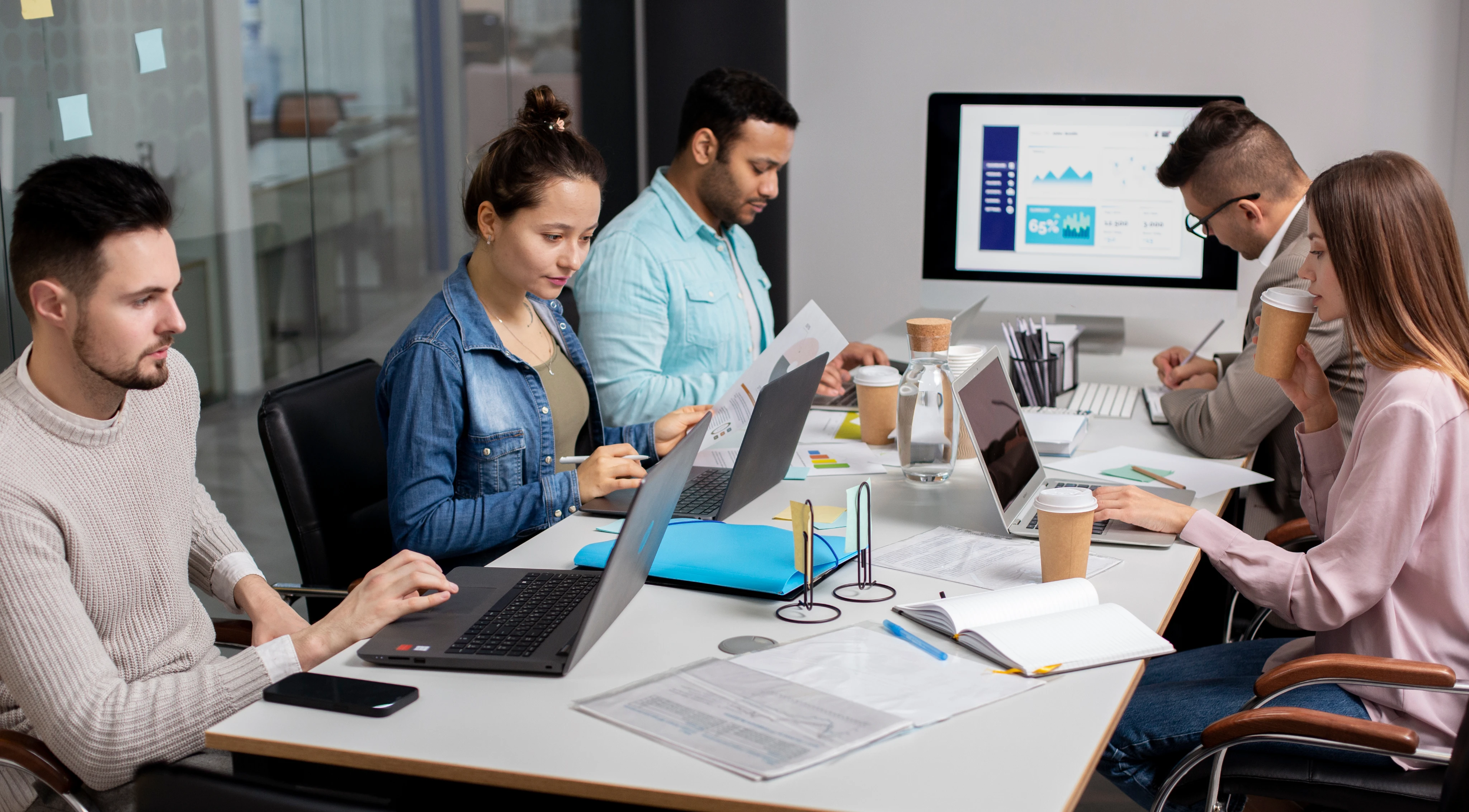 Five young professionals collaborating at a conference table with laptops, documents, coffee cups, and a large monitor displaying charts.