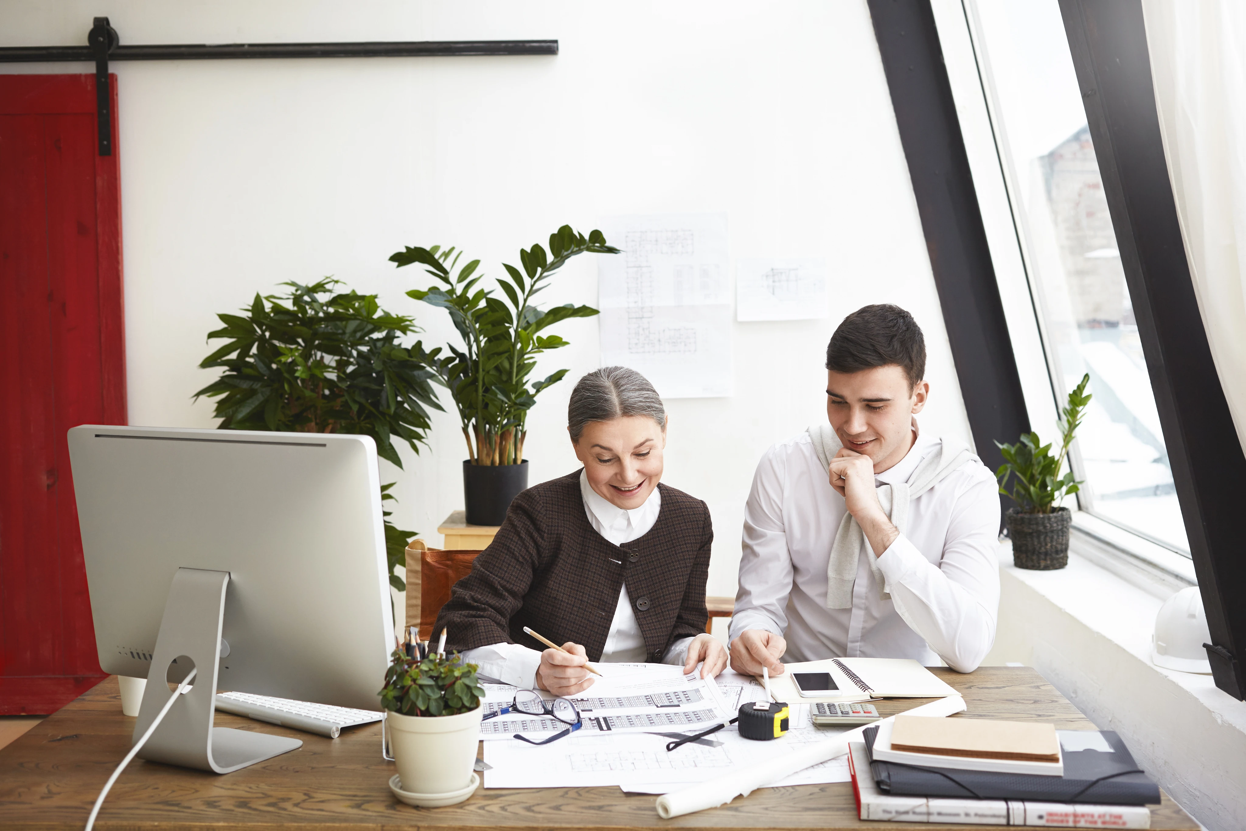 Two colleagues, a woman and a man, reviewing architectural plans and documents together at a desk in a bright office.