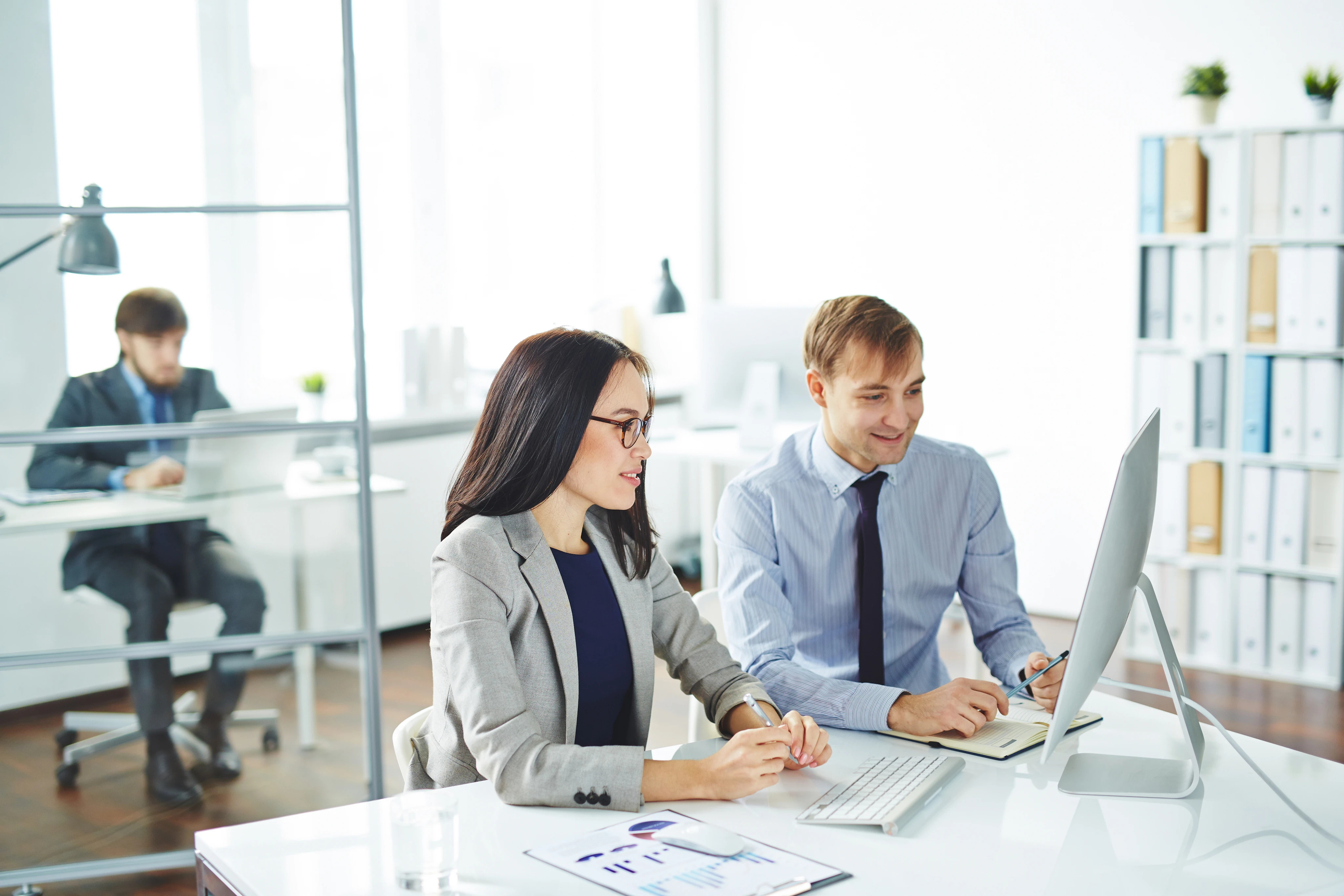 Two business professionals, a man and a woman, working together at a desktop computer in a modern office, with another man working on a laptop in the background.