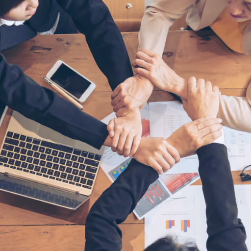 Four people join hands over a table with laptop, phone, and business documents symbolizing teamwork, our mission.