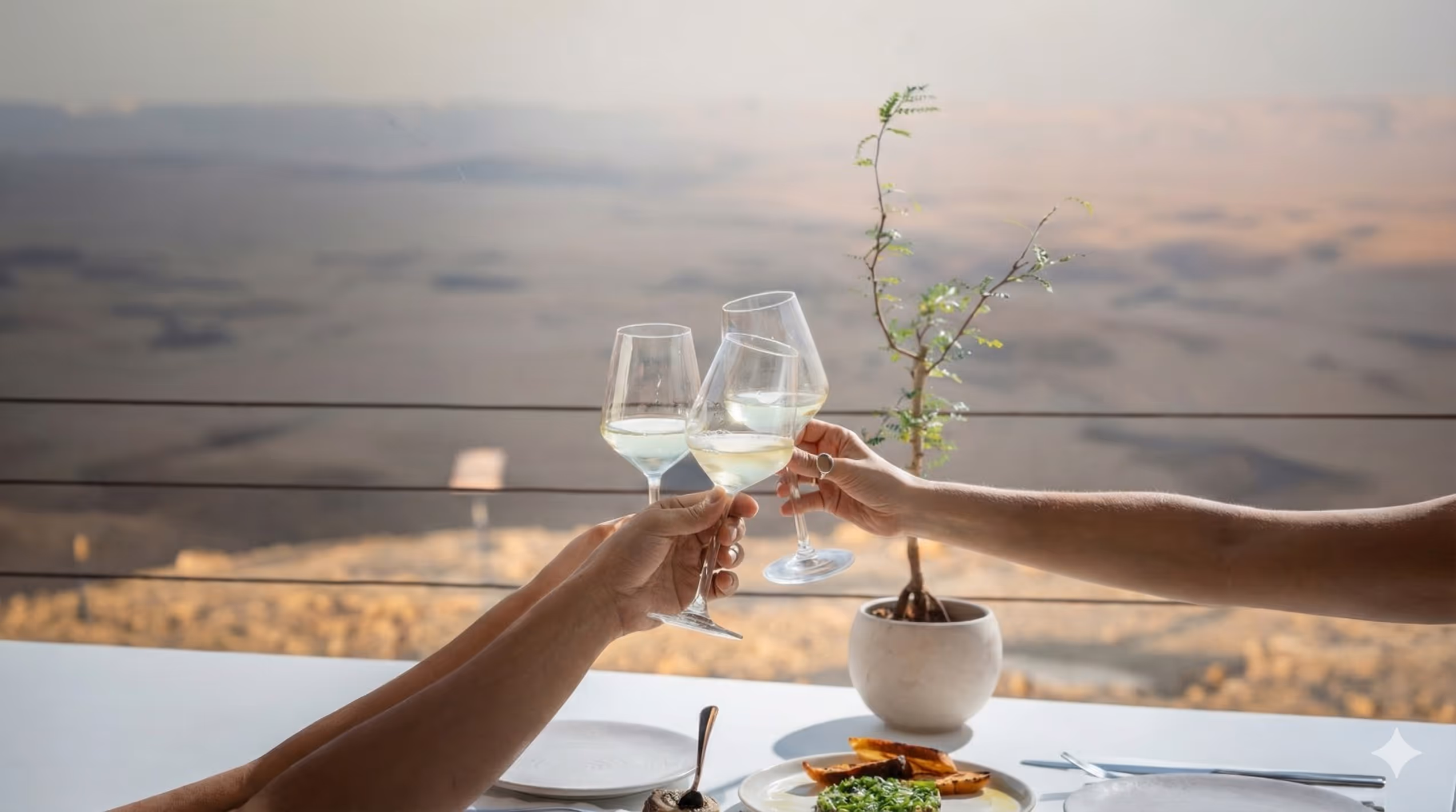 שתי ידיים מוחזקות ומצעצעות כוסות יין לבן על שולחן עם אוכל וצמח בסיר, נוף מדברי ברקע.