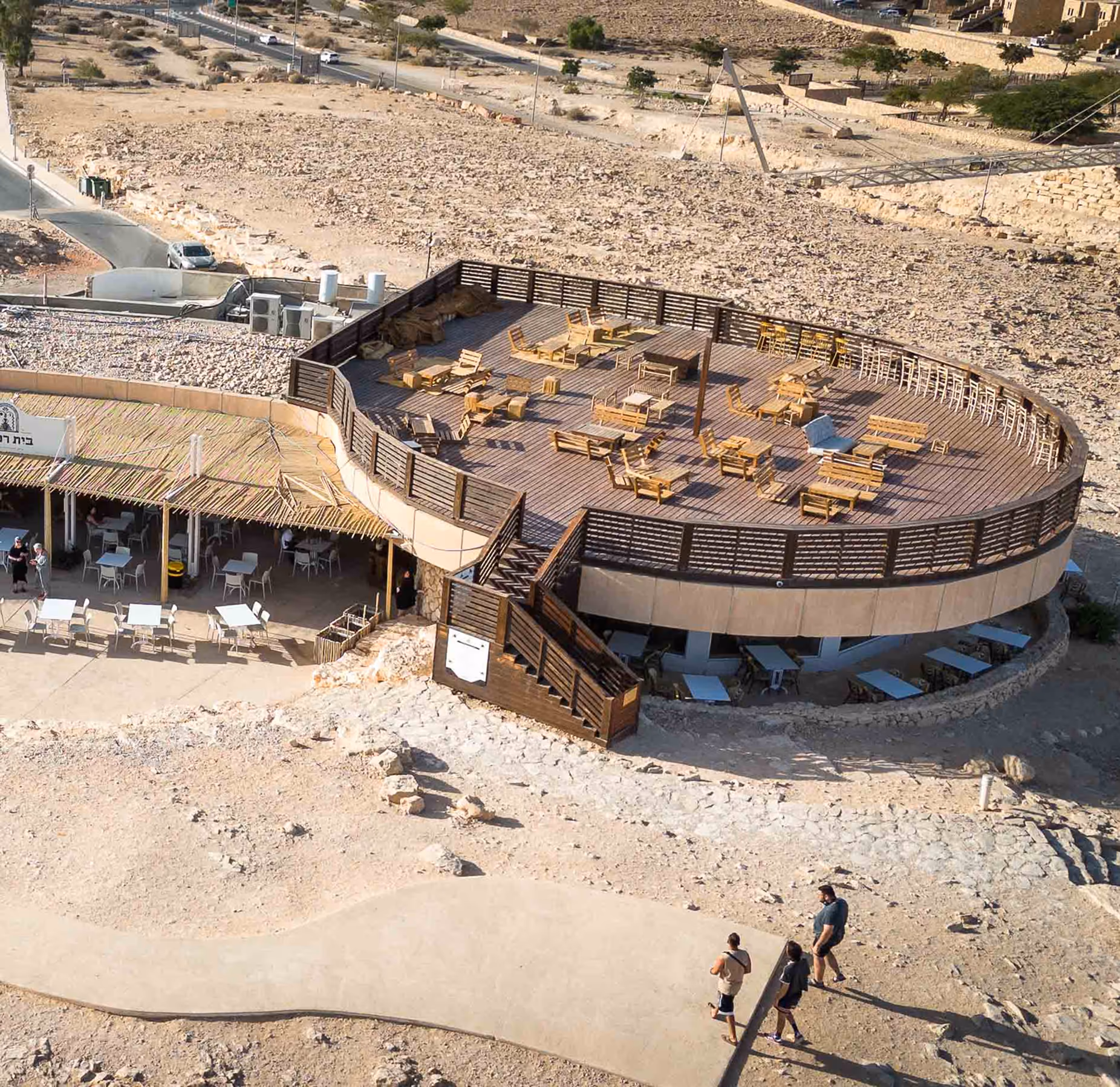 מרפסת עגולה עם ריהוט עץ פתוח המדמה בר בנוף מדברי, עם שביל בטון ואנשים הולכים בסביבה.