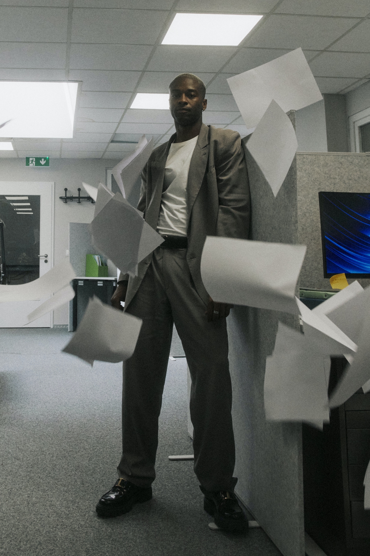 Man in a gray suit stands in an office cubicle with papers flying around him.