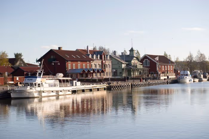 Fartyg vid kaj med röda och gröna byggnader och träd längs en lugn vattenväg under en klar himmel.