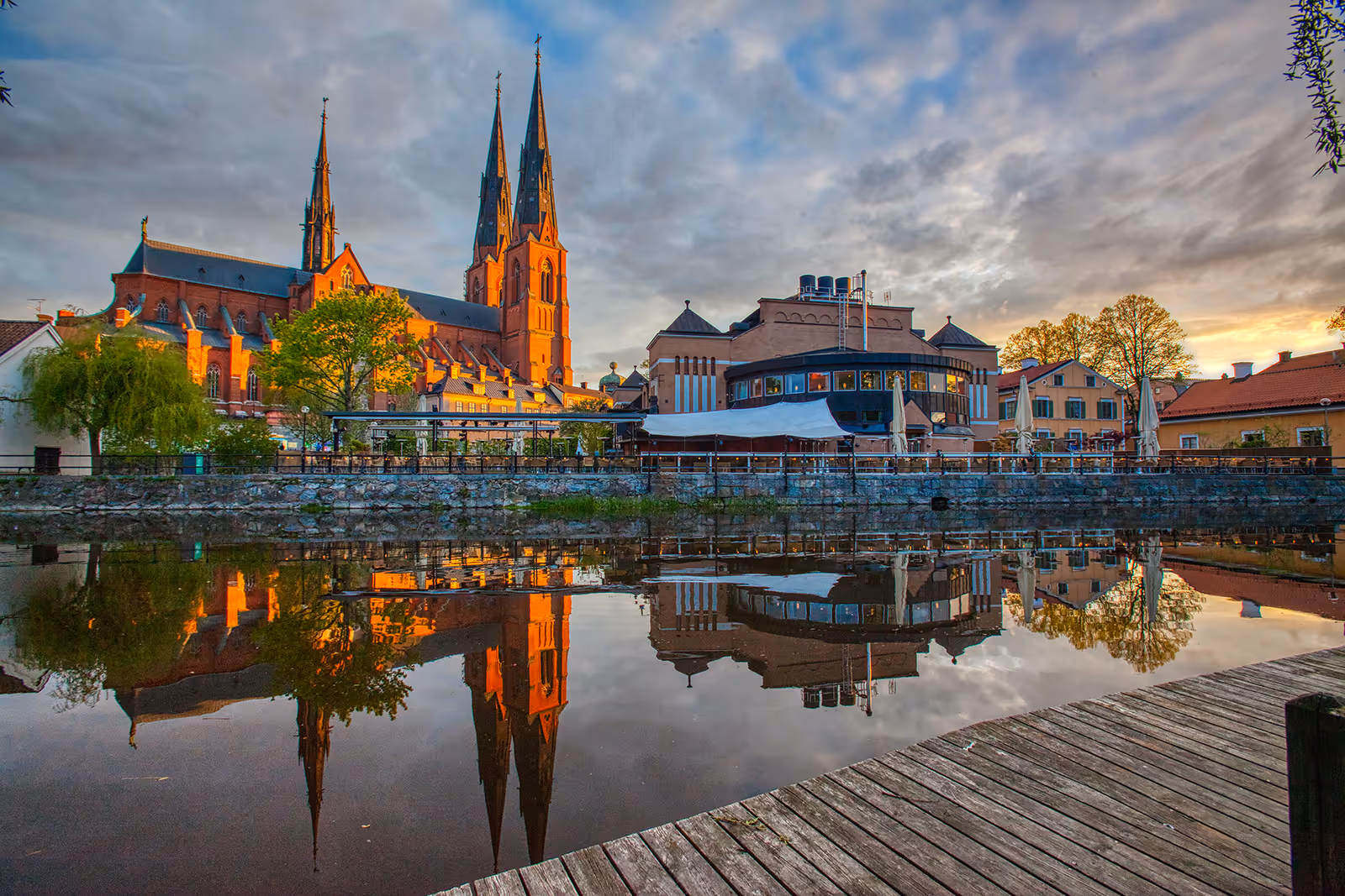 Uppsala domkyrka och omgivande byggnader speglade i en lugn å vid solnedgång.