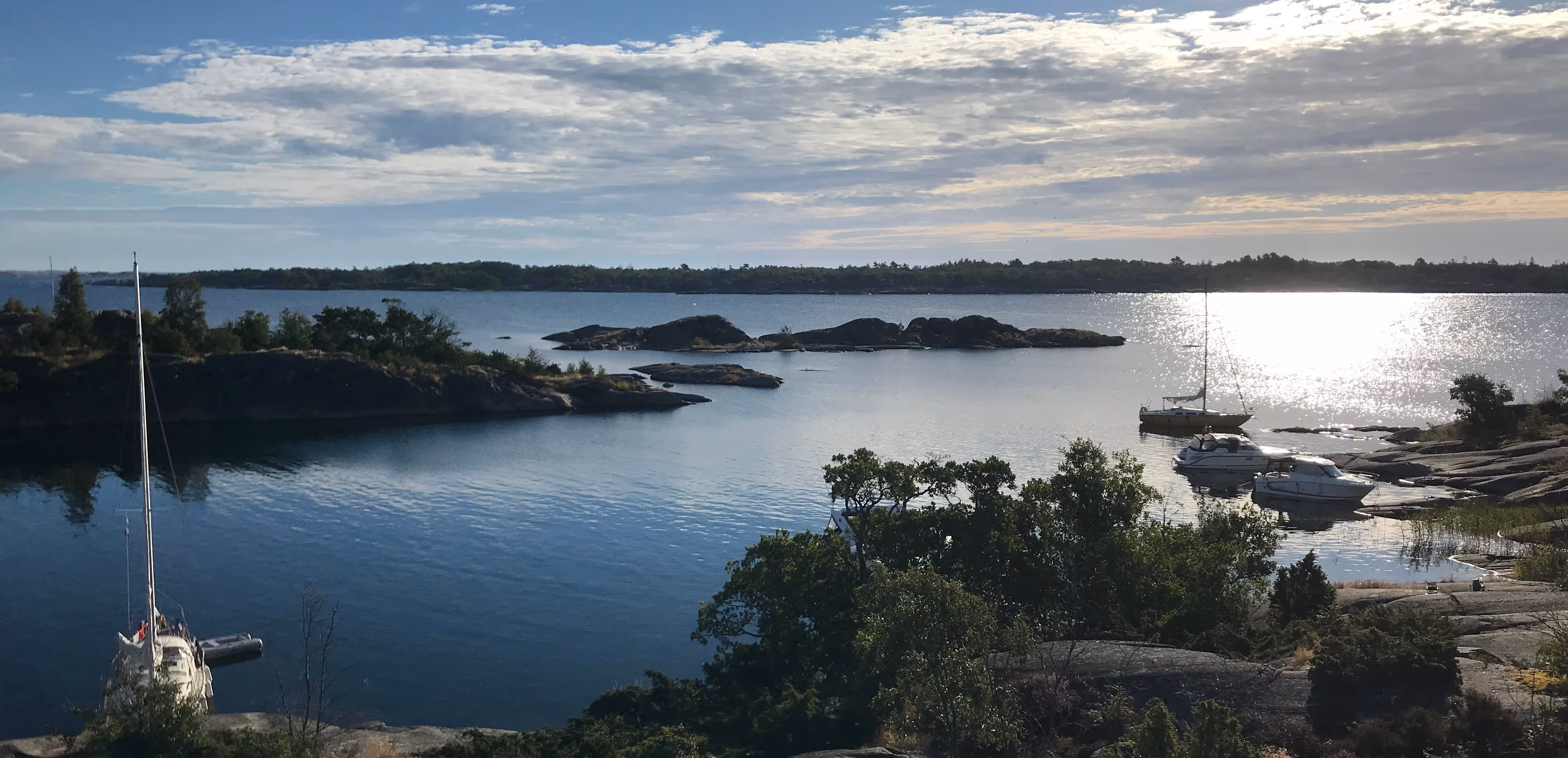 Skärgård med klippor, träd och stilla vatten där flera båtar ligger förtöjda under en molnig himmel.