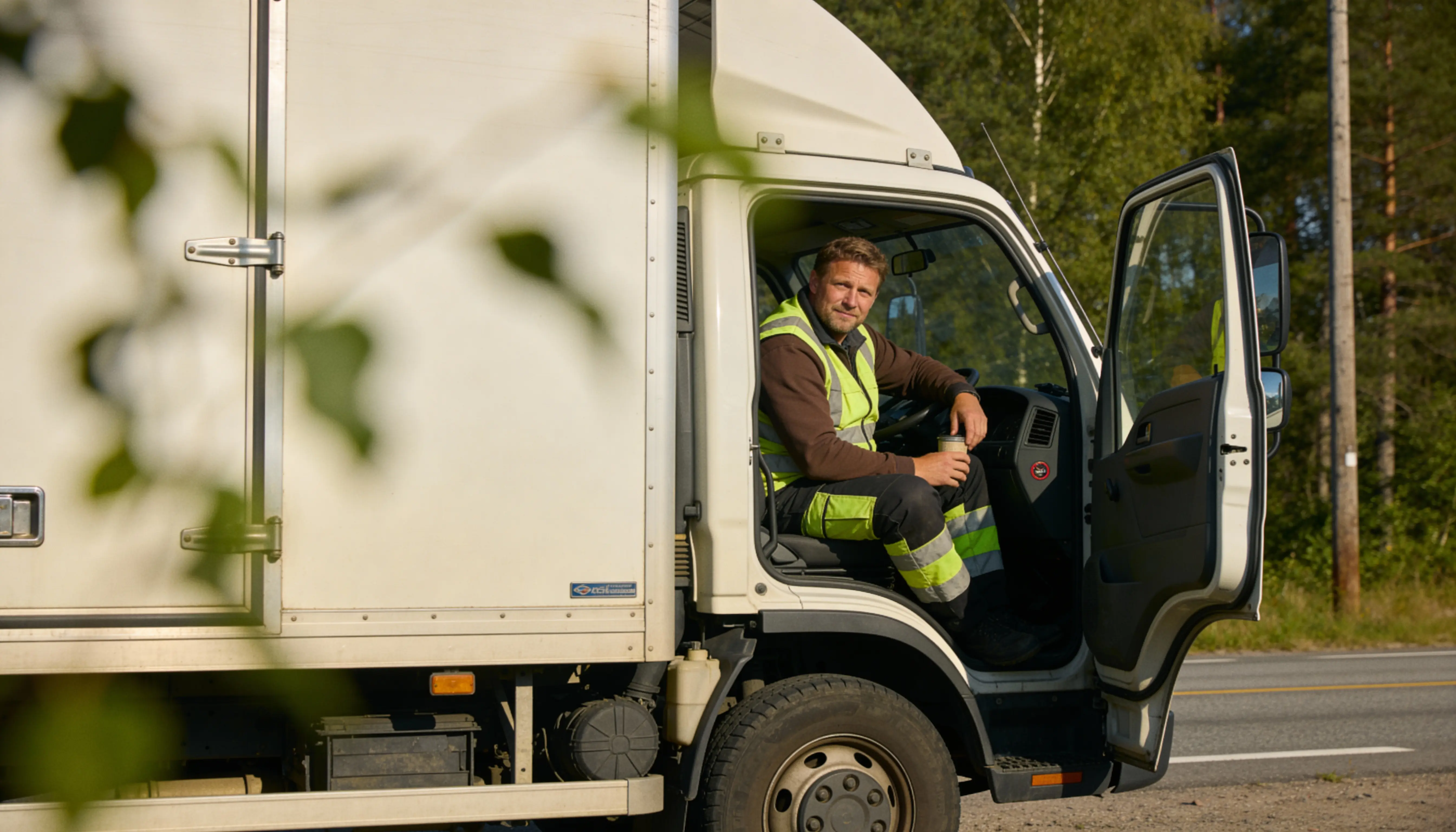 Man in reflective workwear sitting in the driver's seat of a white delivery truck with the door open, holding a cup.