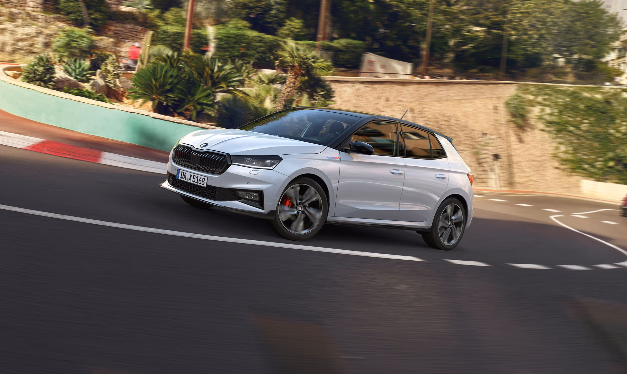 Ein Škoda Fabia 130 in der Lackierung Moon-Weiß Perleffekt fährt auf einer kurvigen Straße, die in eine hügelige, mediterrane Landschaft eingebettet ist. Das Fahrzeug verfügt über das Color Concept Schwarz, erkennbar an Dach, Außenspiegeln und Kühlergrillrahmen in schwarzer Farbe. 