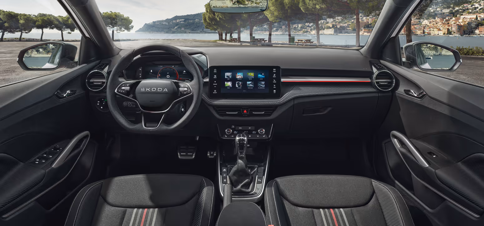 Der Innenraum des Škoda Fabia 130 zeigt das 3-Speichen-Sportmultifunktionslenkrad, das digitale Cockpit sowie das zentrale Infotainment-Display. Die Armaturentafel ist mit einer Dekorleiste in Carbon-Optik und einer roten Akzentlinie versehen. Abgerundet wird das Interieur durch eine Edelstahlpedalerie und den Ausblick durch die Frontscheibe auf eine mediterrane Küstenlandschaft.