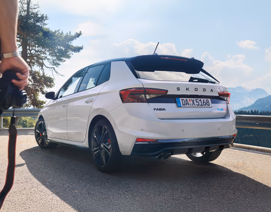 Heckansicht des Škoda Fabia 130 in Moon-Weiß Perleffekt bei Sonnenschein auf einer Bergstraße, wobei im Vordergrund angeschnitten eine Person steht, die eine Fotokamera in der Hand hält. Das Fahrzeug verfügt über das Color Concept Schwarz mit schwarzem Dach und Dachkantenspoiler sowie über sportliche Leichtmetallfelgen. Details wie der Škoda-Schriftzug, das 130-Emblem und der Heckdiffusor mit Doppelendrohr runden das Erscheinungsbild ab.