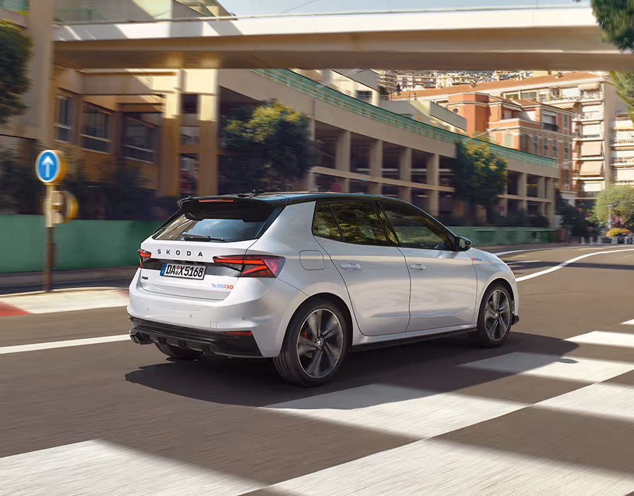 Heckansicht des Škoda Fabia 130 in der Lackierung Moon-Weiß Perleffekt, der dynamisch durch eine mediterrane städtische Kulisse fährt. Das Bild zeigt das schwarze Dach des Color Concept Schwarz sowie den Dachkantenspoiler und die markanten Heckleuchten. Auf der Heckklappe sind der zentrale Škoda Schriftzug und das spezielle 130-Emblem zu erkennen.