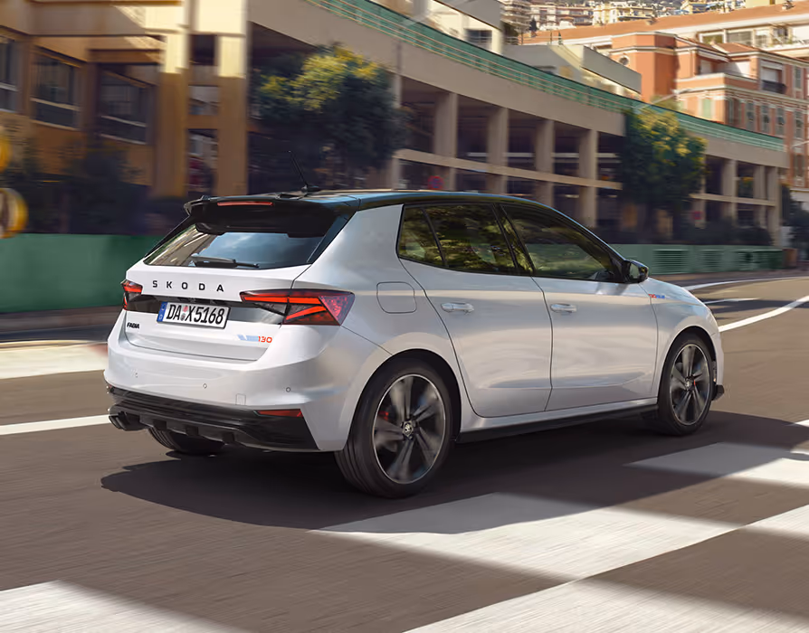 Heckansicht des Škoda Fabia 130 in der Lackierung Moon-Weiß Perleffekt, der dynamisch durch eine mediterrane städtische Kulisse fährt. Das Bild zeigt das schwarze Dach des Color Concept Schwarz sowie den Dachkantenspoiler und die markanten Heckleuchten. Auf der Heckklappe sind der zentrale Škoda Schriftzug und das spezielle 130-Emblem zu erkennen.