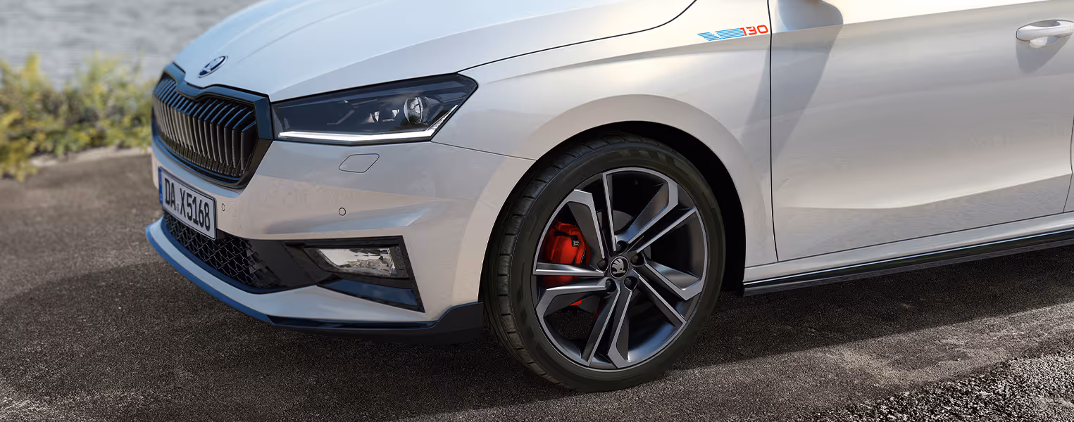 Nahaufnahme der Frontpartie des Škoda Fabia 130 in Moon-Weiß Perleffekt mit schwarzem Kühlergrillrahmen und Außenspiegeln. Besonders deutlich ist das spezielle "130"-Emblem am vorderen Kotflügel oberhalb des Rades zu erkennen. Das Bild zeigt zudem detailliert die sportlichen Leichtmetallfelgen, die den Blick auf die dahinterliegenden roten Bremssättel freigeben.