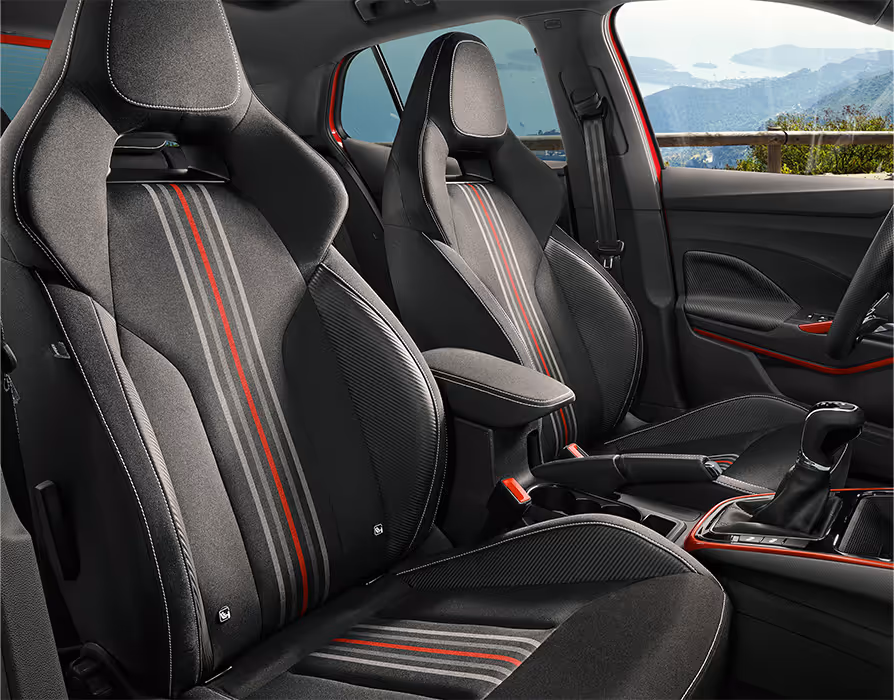 Der Innenraum des Škoda Fabia 130 präsentiert die schwarzen Sportsitze im Monte-Carlo-Design mit integrierten Kopfstützen und Seitenwangen in Carbon-Optik. Charakteristisch sind die vertikalen Zierstreifen in Grau und Rot auf den Sitzmittelbahnen sowie die weißen Kontrastnähte. Durch das Seitenfenster ist im Hintergrund eine mediterrane Küstenlandschaft sichtbar.