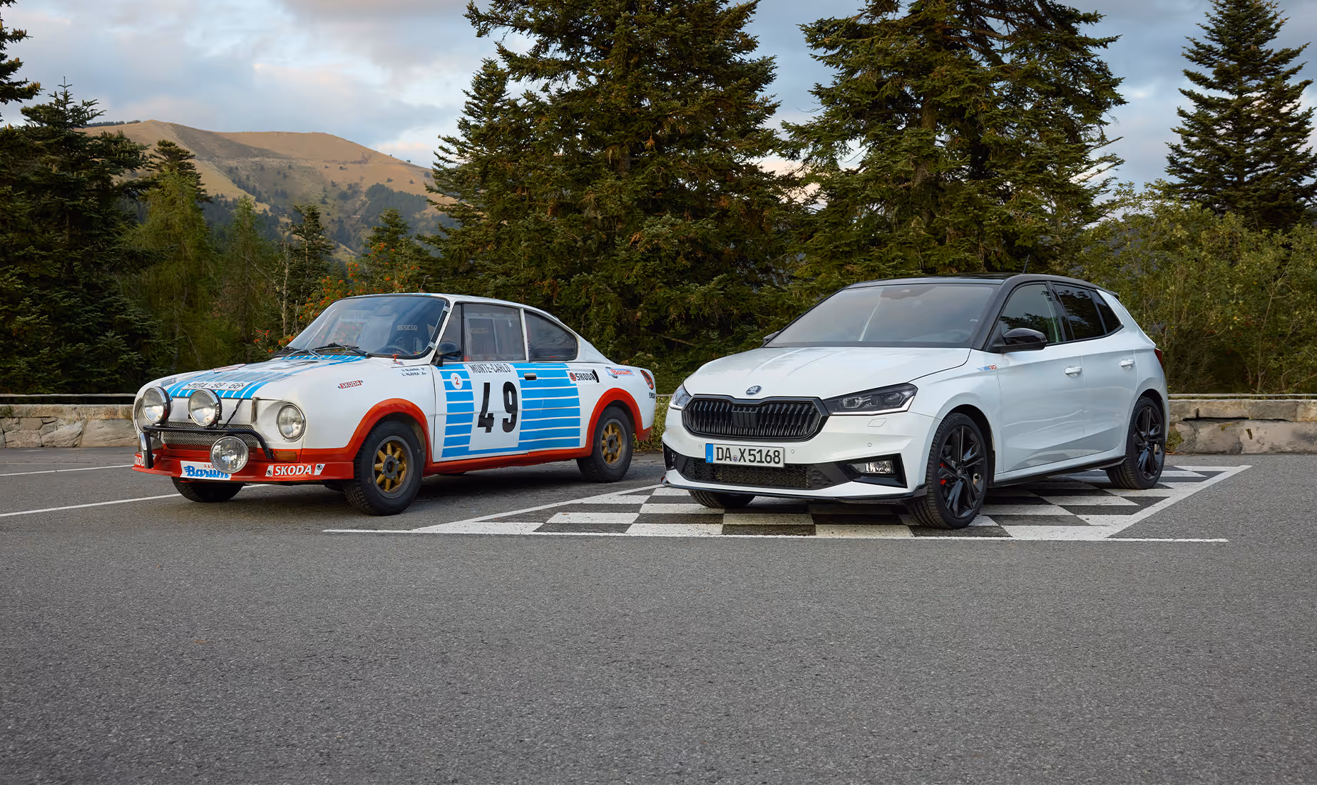 Der neue Škoda Fabia 130 in Moon-Weiß Perleffekt steht rechts neben einem historischen Škoda Rallye-Fahrzeug mit der Startnummer 49 auf einem asphaltiertem Untergrund mit schwarz-weißem Schachbrettmuster. Die beiden Fahrzeuge sind vor einer bewaldeten mediterranen Hügellandschaft positioniert.