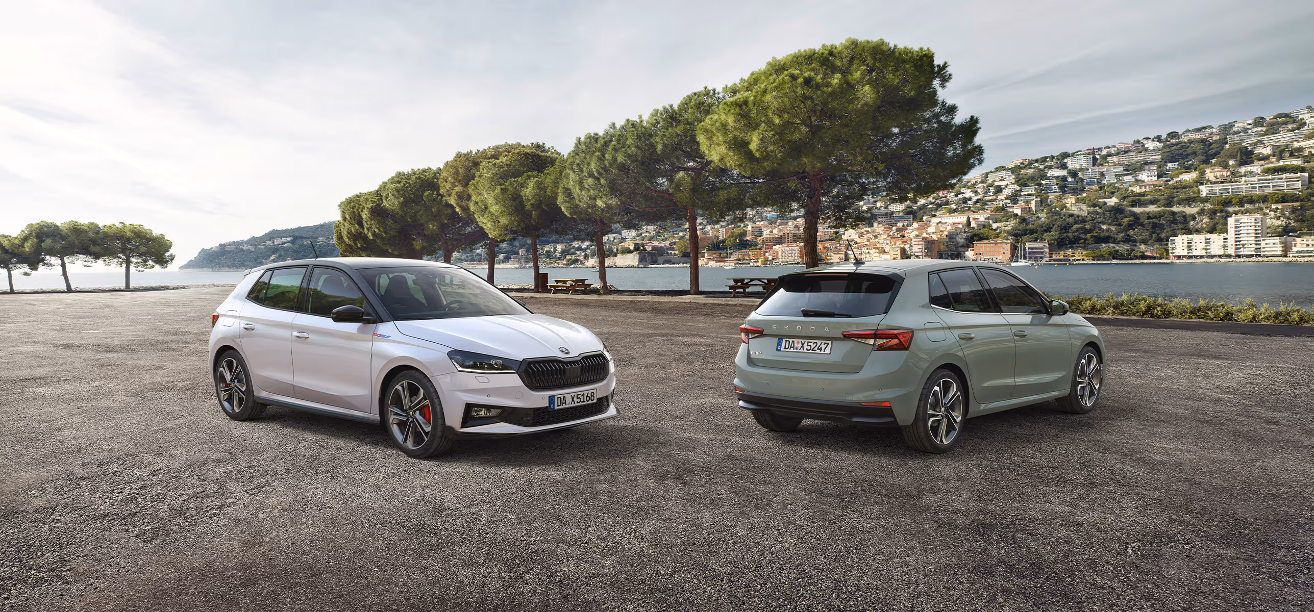 Zwei Škoda Fabia Fahrzeuge stehen nebeneinander vor einer mediterranen Bucht mit einer Stadt am Hang im Hintergrund. Links zeigt sich der Fabia 130 in Moon-Weiß Perleffekt mit schwarzem Dach und markanter Frontpartie. Rechts daneben ist ein Serienmodell in der Lackierung Timiano-Grün in der Heckansicht zu sehen.