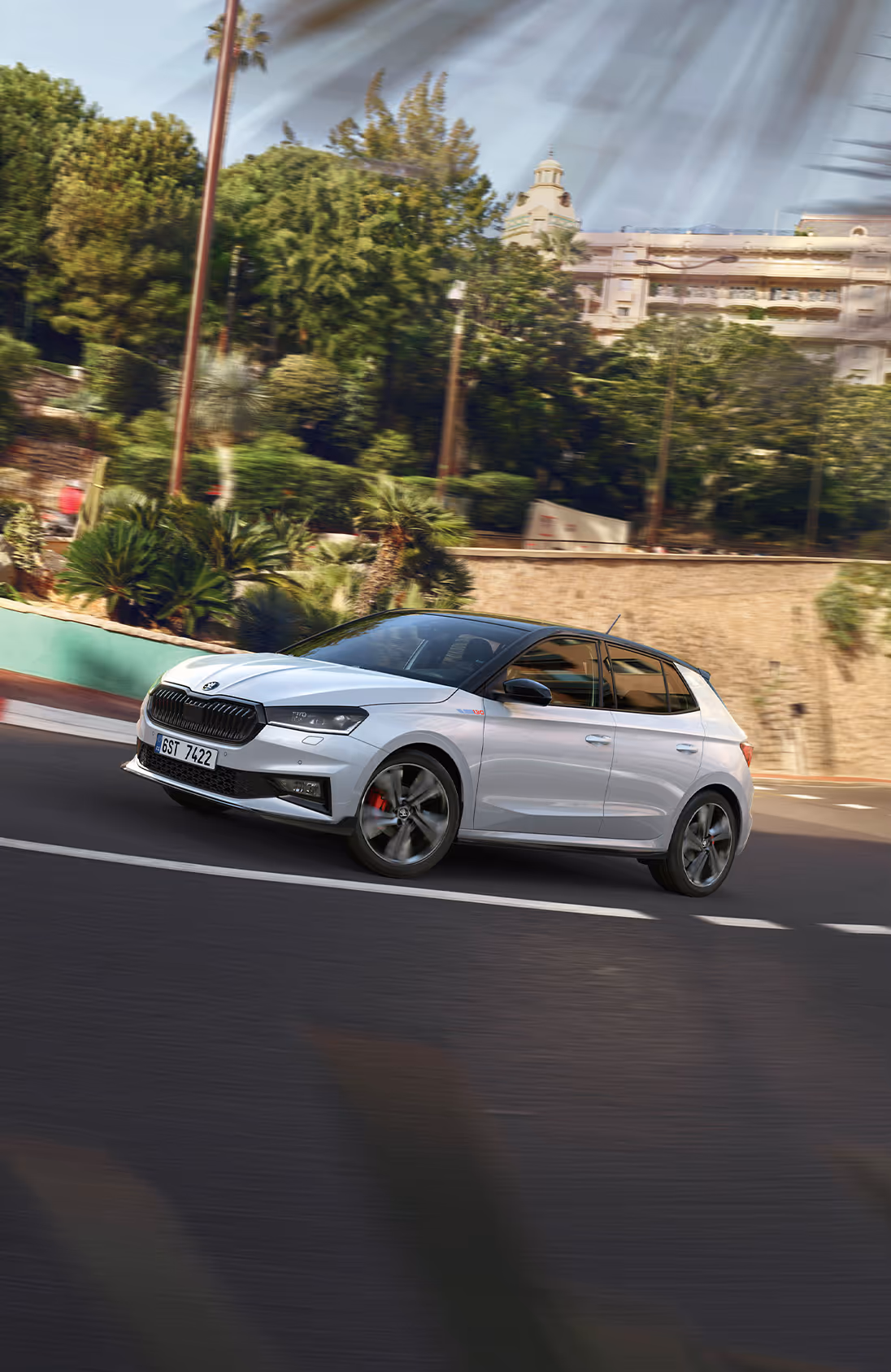 Ein Škoda Fabia 130 in der Lackierung Moon-Weiß Perleffekt fährt auf einer kurvigen Straße, die in eine hügelige, mediterrane Landschaft eingebettet ist. Das Fahrzeug verfügt über das Color Concept Schwarz, erkennbar an Dach, Außenspiegeln und Kühlergrillrahmen in schwarzer Farbe. 