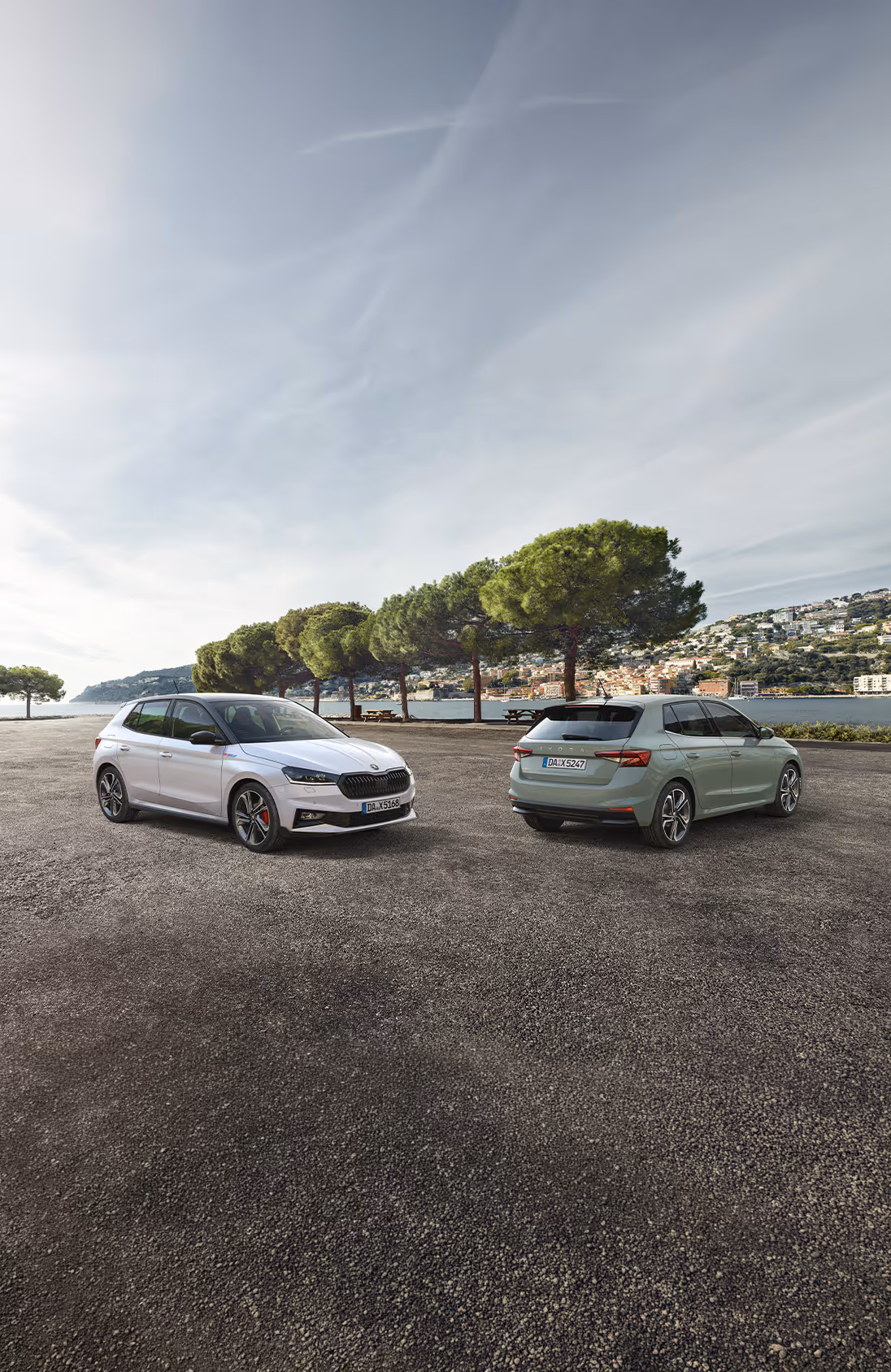 Zwei Škoda Fabia Fahrzeuge stehen nebeneinander vor einer mediterranen Bucht mit einer Stadt am Hang im Hintergrund. Links zeigt sich der Fabia 130 in Moon-Weiß Perleffekt mit schwarzem Dach und markanter Frontpartie. Rechts daneben ist ein Serienmodell in der Lackierung Timiano-Grün in der Heckansicht zu sehen.