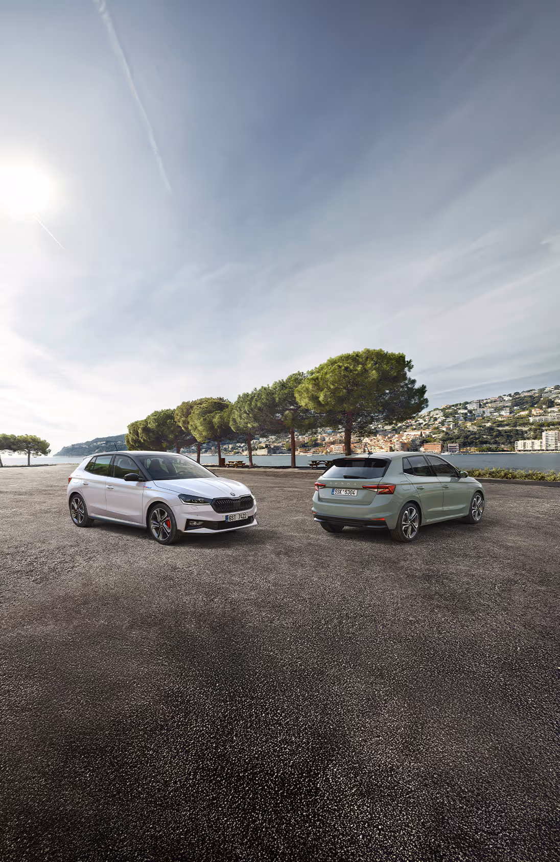Zwei Škoda Fabia Fahrzeuge stehen nebeneinander vor einer mediterranen Bucht mit einer Stadt am Hang im Hintergrund. Links zeigt sich der Fabia 130 in Moon-Weiß Perleffekt mit schwarzem Dach und markanter Frontpartie. Rechts daneben ist ein Serienmodell in der Lackierung Timiano-Grün in der Heckansicht zu sehen.