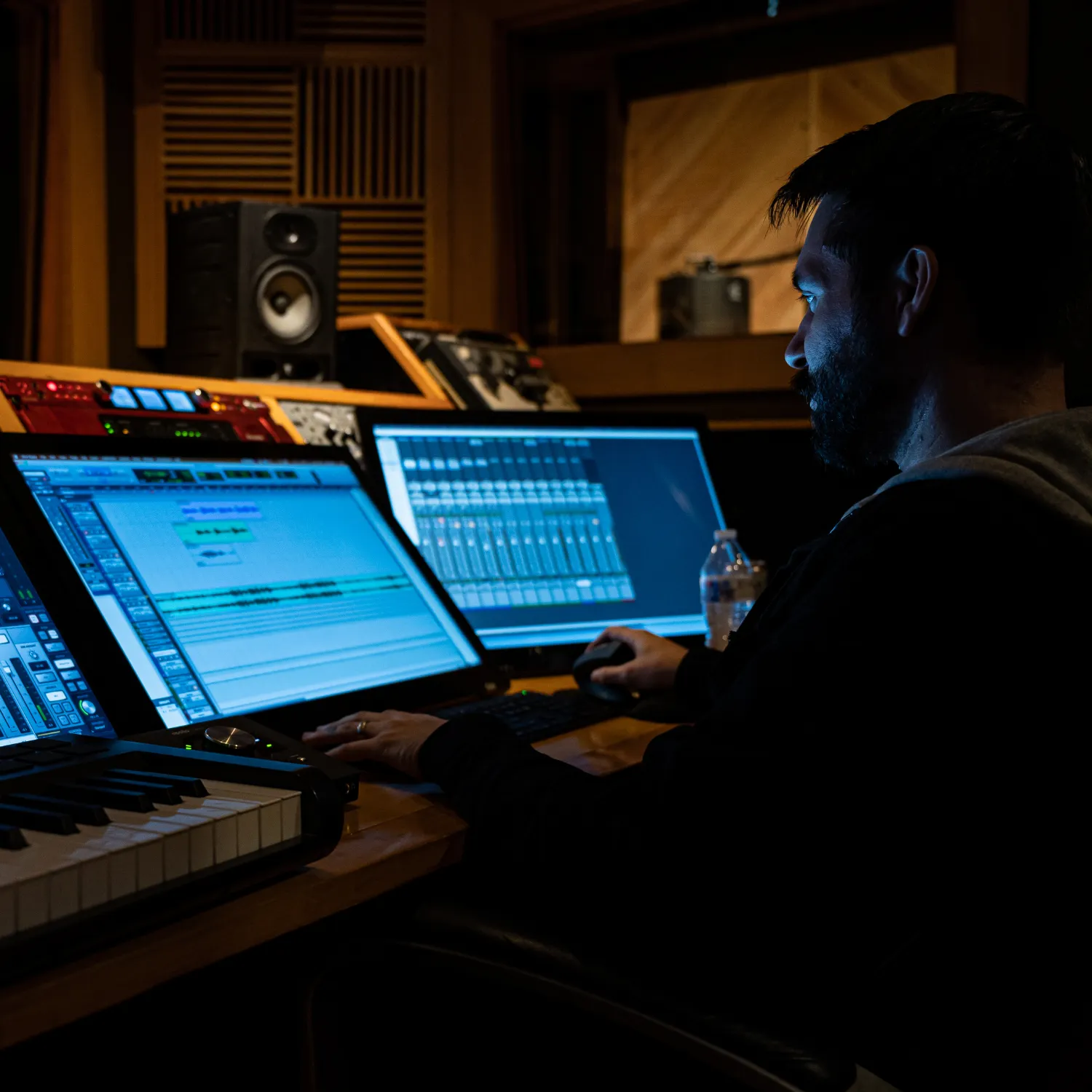 Man with a beard working on audio editing software at a recording studio desk with multiple monitors and a keyboard.